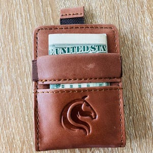 May include: A brown leather card holder featuring a debossed horse head logo on the front pocket. The wallet includes a pull tab at the top and a side elastic band, with a United States banknote partially visible in the main storage compartment.