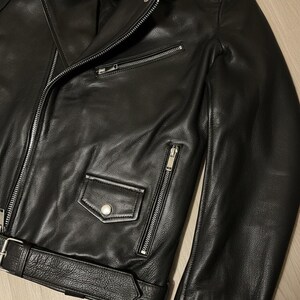 May include: A black leather jacket with a classic biker style. Features include a notched lapel, asymmetrical zipper closure, multiple zippered pockets, and a belted waist. The jacket is made of genuine leather.