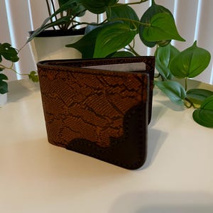 Puede incluir: Cartera de cuero marrón con un estampado de piel de serpiente en la parte delantera. La cartera está abierta, revelando un vistazo del interior. Los bordes y las esquinas son de un marrón más oscuro. La cartera está sobre una superficie blanca.