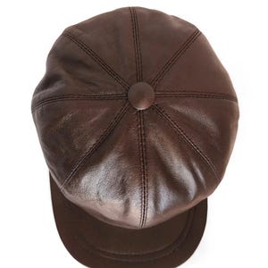 Puede incluir: Una gorra de estilo boina en cuero marrón oscuro con una construcción de ocho paneles, costuras visibles a lo largo de las uniones, un botón central forrado en el centro y una visera corta y curva en la parte frontal de la gorra.