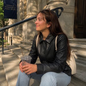 May include: A black leather jacket with a button-up front. The jacket is worn over a light-colored top and paired with light blue jeans. A clear plastic cup with a straw is held in the hands. A white backpack is on the back. The background includes a building with a banner that reads "SCHOOL OF INTERNATIONAL PUBLIC AFFAIRS."