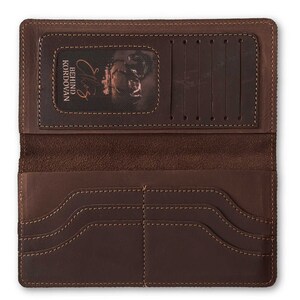 May include: Open, brown leather wallet with multiple card slots and compartments. The wallet features visible stitching and a textured surface. The interior includes a clear window with the text "BEHIND KORDOVAN" and a signature.
