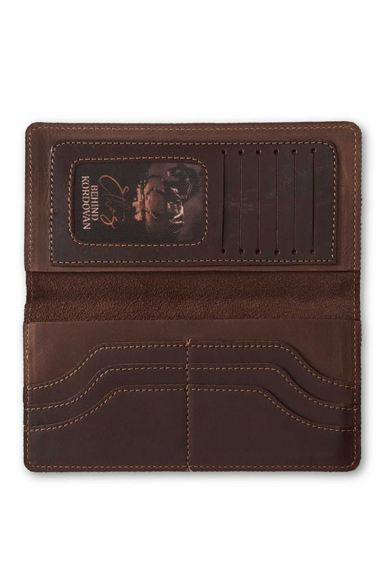 May include: Open, brown leather wallet with multiple card slots and compartments. The wallet features visible stitching and a textured surface. The interior includes a clear window with the text "BEHIND KORDOVAN" and a signature.