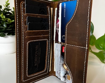 May include: A brown leather bifold wallet featuring white contrast stitching, multiple vertical card slots on the left side, a clear ID window with a KORDOVAN logo, and several horizontal pockets on the right side containing various bank cards.
