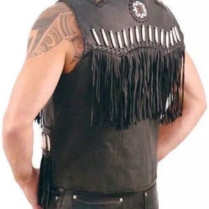 May include: Black leather vest with fringe and decorative beading. The vest features white and tan beads along the shoulders and back, with long black fringe along the bottom. The vest is sleeveless and has a classic biker style.