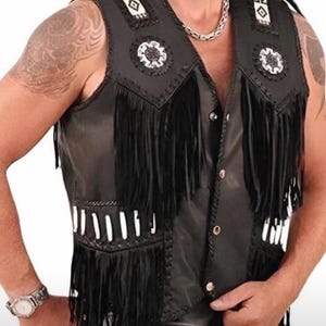 May include: Black leather vest with fringe detailing. The vest features decorative white and black accents on the shoulders and chest, along with white beadwork. The vest has a button-up front and a classic biker style.