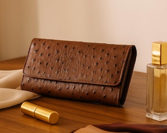 May include: A brown leather wallet with a textured surface and a flap closure. A gold-colored perfume bottle and a small gold tube are also on the wooden surface. A white cloth is draped on the left.