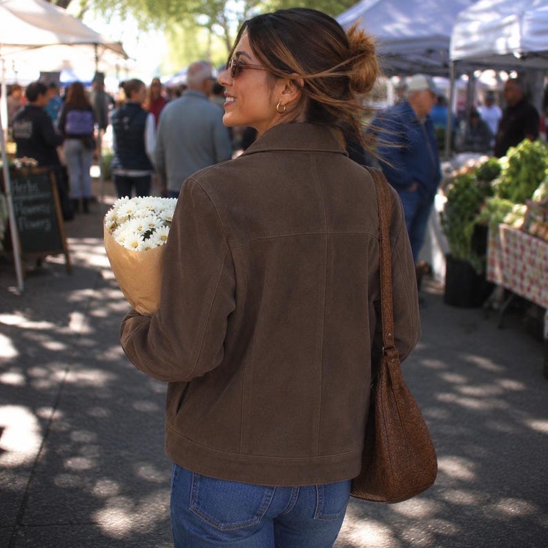 May include: A brown suede jacket with a collared neckline and a matching brown shoulder bag. The person is holding a bouquet of white flowers wrapped in brown paper. The person is wearing blue jeans and sunglasses.
