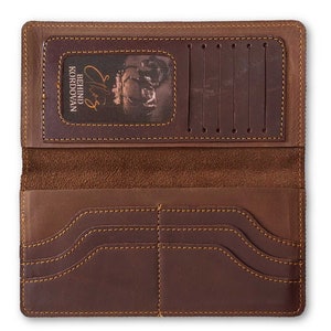 May include: Open brown leather wallet with multiple card slots and bill compartments. The wallet features a clear ID window and is stitched with a contrasting thread. The text "BEHIND KORDOVAN" is visible on the ID window.