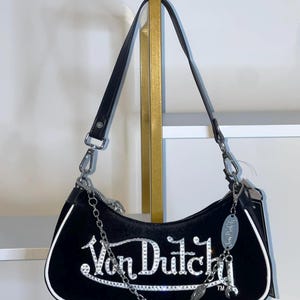 以下が含まれることがあります： 黒いベルベットのショルダーバッグ。シルバーのラインストーンで「Von Dutch」の文字が施されています。白いトリム、黒いレザーストラップ、シルバーのチェーンディテールが特徴です。シルバーのチャームがジッパーからぶら下がっています。