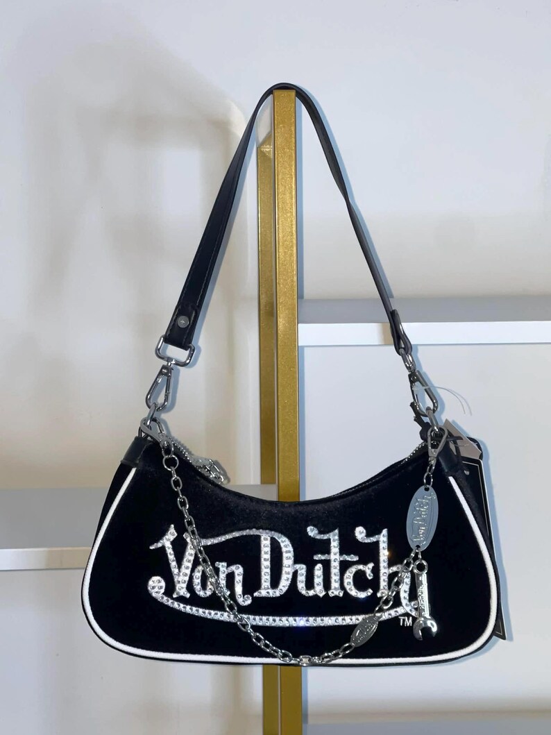 以下が含まれることがあります： 黒いベルベットのショルダーバッグ。シルバーのラインストーンで「Von Dutch」の文字が施されています。白いトリム、黒いレザーストラップ、シルバーのチェーンディテールが特徴です。シルバーのチャームがジッパーからぶら下がっています。