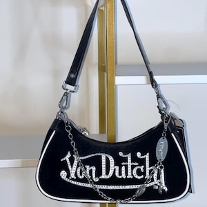 以下が含まれることがあります： 白いラインストーンで「Von Dutch」の文字が入った黒いショルダーバッグ。バッグは白いトリム、シルバーチェーン、黒いストラップが付いています。シルバーのチャームが片側からぶら下がっています。
