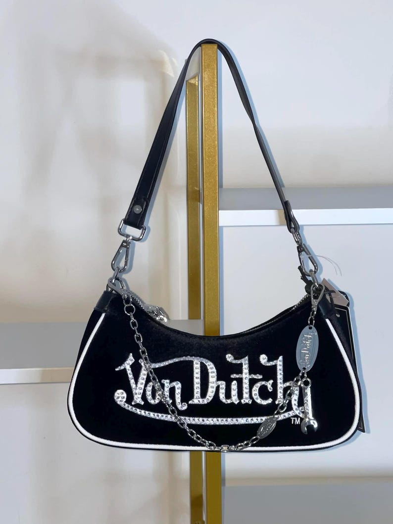 以下が含まれることがあります： 白いラインストーンで「Von Dutch」の文字が入った黒いショルダーバッグ。バッグは白いトリム、シルバーチェーン、黒いストラップが付いています。シルバーのチャームが片側からぶら下がっています。
