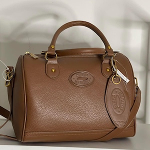 May include: Brown leather satchel bag with a top handle and a detachable shoulder strap. The bag features gold-tone hardware, a zip closure, and the embossed brand name "STEVE MADDEN". A matching leather tag hangs from the handle.