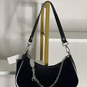 以下が含まれることがあります： 白のトリムとシルバーチェーンストラップが付いた黒のショルダーバッグ。バッグには短いハンドルと装飾的なシルバーチャームが付いています。バッグは柔らかくビロードのような素材で作られています。