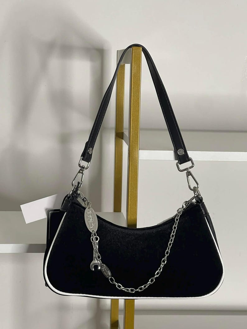 以下が含まれることがあります： 白のトリムとシルバーチェーンストラップが付いた黒のショルダーバッグ。バッグには短いハンドルと装飾的なシルバーチャームが付いています。バッグは柔らかくビロードのような素材で作られています。