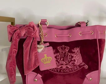 May include: A deep magenta velvet handbag with pink leather trim, featuring a large velvet bow on the side, gold-tone hardware, and a gold chain charm with a heart and a pink tag displaying a gold crown logo and the text Juicy Couture.