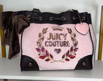May include: A light pink velour handbag with dark brown faux leather trim, featuring a large brown bow on the side. The front displays the text JUICY COUTURE in gold sequins surrounded by a sequined wreath and crown, with a pink heart-shaped charm.