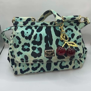 May include: A small handbag with a teal and black leopard print pattern. The bag has a top handle and a detachable shoulder strap. It features gold-tone hardware and a cherry charm. A small metal plaque with text is on the front.