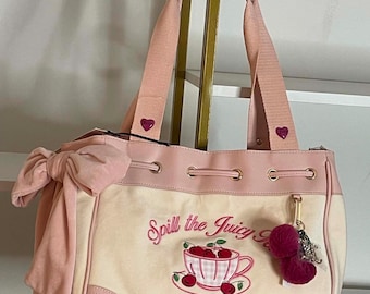 May include: A cream and light pink tote bag featuring a large side bow, two shoulder straps with small heart accents, and a drawstring top. The front displays an embroidered teacup with cherries and the text Spill the Juicy and Juicy Couture.