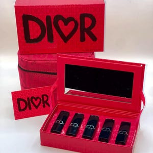 May include: A red gift set featuring lipsticks and a mirror. The set includes a box with the text "DIOR" and a heart symbol, a small round case, and a card with the same text. The lipsticks are in black cases.