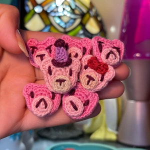 May include: A collection of handmade crochet bear face appliques in various shades of pink. Some bears have a red bow, a purple party hat, or no accessories. The bears have brown stitched details for the eyes, nose, and mouth.