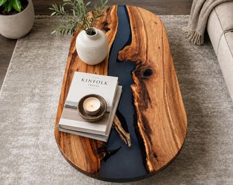 May include: An oval coffee table featuring a natural wood slab with a dark gray resin river running through the center. The table surface holds a white ceramic vase with green foliage, a stack of Kinfolk Travel books, and a small candle.