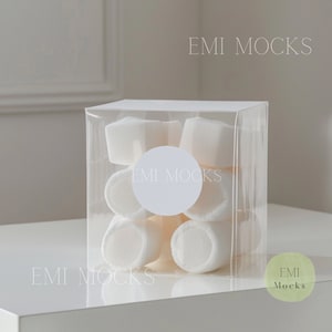 Puede incluir: Una caja de plástico transparente llena de malvaviscos blancos. La caja tiene una pegatina circular blanca con el texto "EMI MOCKS" en ella.