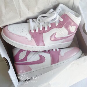 Puede incluir: Un par de zapatillas personalizadas blancas y rosas con un logotipo Swoosh rosa. Las zapatillas est&aacute;n sobre una superficie blanca con el texto "EMI'S CUSTOMS" y "SNEAKERS & SHOES" impreso en la superficie.