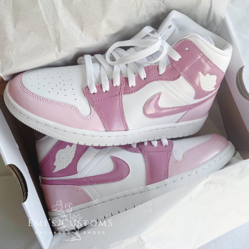 Puede incluir: Un par de zapatillas personalizadas blancas y rosas con un logotipo Swoosh rosa. Las zapatillas est&aacute;n sobre una superficie blanca con el texto "EMI'S CUSTOMS" y "SNEAKERS & SHOES" impreso en la superficie.