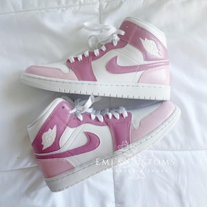 Puede incluir: Un par de zapatillas blancas de ca&ntilde;a alta con detalles en rosa y rosa claro. Las zapatillas tienen un logotipo swoosh blanco y una suela rosa. El texto "EMI'S CUSTOMS" es visible en la parte inferior de la imagen.