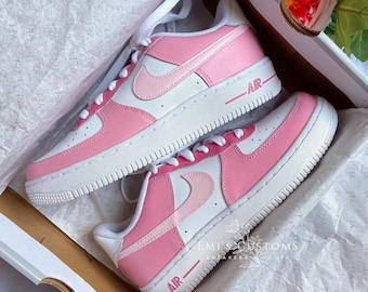 Puede incluir: Un par de zapatillas Nike Air Force 1 personalizadas, blancas y rosas, con un logotipo Swoosh rosa y la palabra "AIR" en el lateral. Las zapatillas están en una caja de zapatos blanca con una flor rosa en la parte superior.