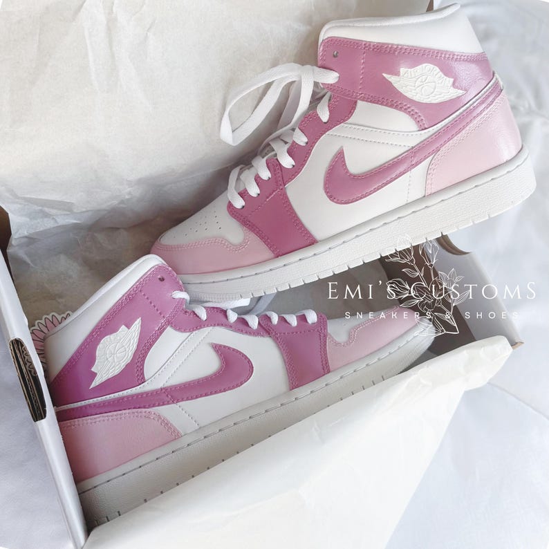 Puede incluir: Un par de zapatillas blancas y rosas de ca&ntilde;a alta con un logotipo Swoosh rosa. Las zapatillas est&aacute;n en una caja blanca con el texto "EMI'S CUSTOMS" y "SNEAKERS & SHOES" impreso en ella.