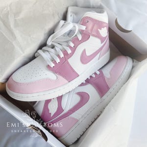 Puede incluir: Un par de zapatillas blancas y rosas de ca&ntilde;a alta con un logotipo Swoosh rosa. Las zapatillas est&aacute;n en una caja blanca con el texto "EMI'S CUSTOMS" y "SNEAKERS & SHOES".