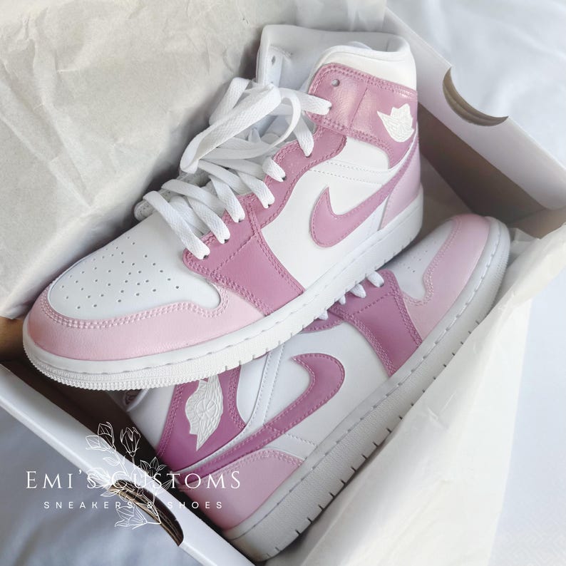 Puede incluir: Un par de zapatillas blancas y rosas de ca&ntilde;a alta con un logotipo Swoosh rosa. Las zapatillas est&aacute;n en una caja blanca con el texto "EMI'S CUSTOMS" y "SNEAKERS & SHOES".