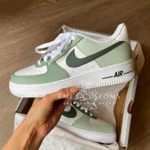 Peut inclure: Une paire de baskets Air Force 1 personnalis&eacute;es dans une bo&icirc;te. Les chaussures sont blanches, vert clair et vert fonc&eacute; avec des lacets blancs et une semelle blanche. Le mot "AIR" est imprim&eacute; sur le c&ocirc;t&eacute;. La bo&icirc;te porte le texte "EMI'S CUSTOMS SNEAKERS & SHOES".