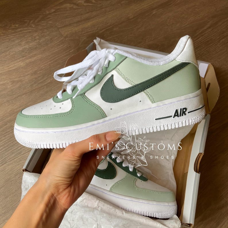 Peut inclure: Une paire de baskets Air Force 1 personnalis&eacute;es dans une bo&icirc;te. Les chaussures sont blanches, vert clair et vert fonc&eacute; avec des lacets blancs et une semelle blanche. Le mot "AIR" est imprim&eacute; sur le c&ocirc;t&eacute;. La bo&icirc;te porte le texte "EMI'S CUSTOMS SNEAKERS & SHOES".