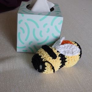 Puede incluir: Un juguete de abeja de crochet con rayas negras y amarillas, alas blancas y detalles naranjas. La abeja está tumbada sobre una superficie marrón claro junto a una caja de pañuelos con un patrón turquesa y blanco.