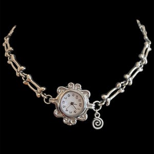 Può includere: Collana orologio color argento con quadrante bianco e numeri neri. L'orologio è circondato da una cornice decorativa e collegato a una catena di maglie a forma di osso. Un piccolo ciondolo a spirale pende dall'orologio.