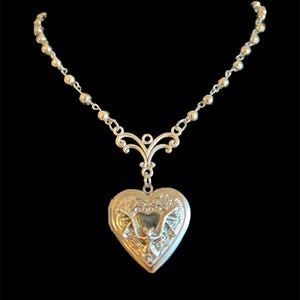 Peut inclure: Collier avec pendentif en forme de cœur argenté. Le médaillon est orné de motifs floraux et décoratifs détaillés. La chaîne du collier est composée de petites perles rondes. Le fond est noir.