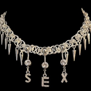 Könnte beinhalten: Silberfarbene Halskette im Choker-Stil mit Kettenmuster. Die Halskette hat hängende, kegelförmige Anhänger und drei Anhänger mit dem Wort "SEX" in glitzernden Buchstaben. Der Hintergrund ist schwarz.