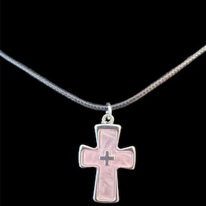 Puede incluir: Collar con colgante de cruz plateada, con centro rosa claro y diseño de cruz más pequeña. El colgante cuelga de un cordón gris sobre un fondo negro.