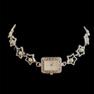 Puede incluir: Un reloj plateado con una esfera rectangular y una correa decorativa en forma de estrella. La esfera del reloj tiene un fondo blanco con números y manecillas negras. La correa presenta eslabones en forma de estrella con una pequeña esfera en el centro.