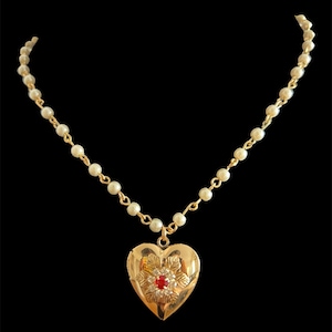 Könnte beinhalten: Goldfarbene herzförmige Medaillon-Halskette mit einer Perlen- und Goldkette. Das Medaillon hat ein Blumendesign mit einem roten Edelstein in der Mitte. Die Halskette wird vor einem schwarzen Hintergrund präsentiert.