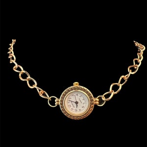 Könnte beinhalten: Eine goldfarbene Halskettenuhr mit rundem Zifferblatt und weißem Zifferblatt. Das Zifferblatt hat schwarze Ziffern und Zeiger. Die Kette besteht aus ineinandergreifenden ovalen Gliedern. Die Uhr ist vor schwarzem Hintergrund.