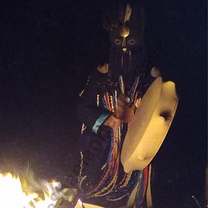Puede incluir: Una persona con un tocado y vestimenta tradicional nativa americana golpea un tambor frente a un fuego. El tambor está hecho de madera y cuero y tiene un diseño colorido. La persona lleva una máscara negra con ojos blancos y una boca roja.