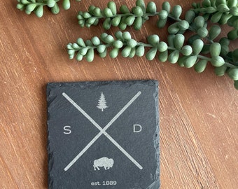 May include: Black slate coaster with a white design. The design features a stylized pine tree, a buffalo silhouette, and the letters "S" and "D" crossed over each other. The text "est. 1889" is below the design.