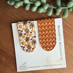 May include: Two magnetic bookmarks attached to a white display card labeled MAGNETIC BOOKMARKS and HANDMADE IN SOUTH DAKOTA. One bookmark features a white background with a floral pattern in yellow and brown, while the other has a brown woven texture.