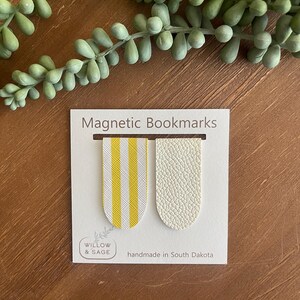 May include: Two magnetic bookmarks, one with yellow and white stripes and the other with a white textured surface. The bookmarks are displayed on a white card with the text "Magnetic Bookmarks", "Willow & Sage", and "handmade in South Dakota".