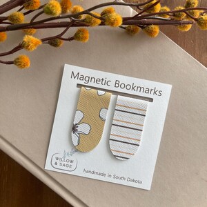 May include: Two magnetic bookmarks on a card with the text "Magnetic Bookmarks" and "Willow & Sage". One bookmark is mustard yellow with a white floral design, the other is white with brown and black stripes. The card reads "handmade in South Dakota".